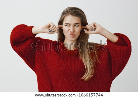 Portrait of distracted uncomfortable charming caucasian girl in loose sweater, covering ears with index fingers, looking left and smirking from dislike, hearing loud awful noise over gray background