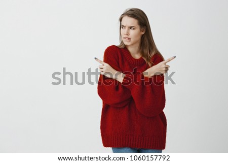 Troubled girl making tough deicison. Doubtful attractive woman in loose red sweater, crossing hands and pointing in different directions, biting lip nervously, wanting something over gray wall