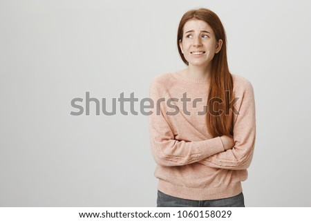 Indoor portrait of cute ginger girl standing with crossed hands on belly, feeling awkward or suffering from pain while looking aside, standing against gray background. Woman has stomachache Royalty-Free Stock Photo #1060158029