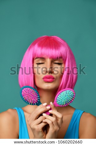 Closeup portrait of happy fashion brunette woman hold two small pink blue yellow small hair comb brush kissing sign in pink wig on modern mint background