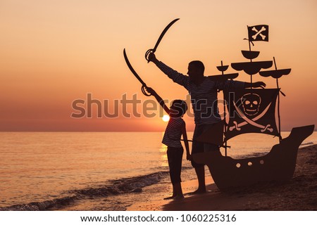 Father and son playing on the beach at the sunset time. They playing with a cardboard pirate ship. People having fun outdoors. Concept of summer vacation and friendly family.
