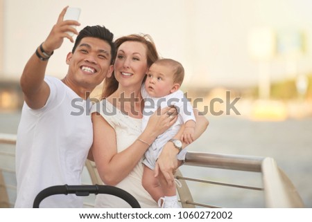 Mixed-race family of three taking selfie on smartphone while standing at riverwalk, cute baby looking at device with curiosity