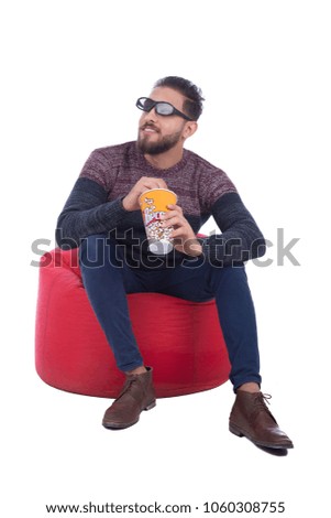 Handsome young man sitting on a bean bag wearing a 3D glasses and holding a popcorn cup in his hand eating it, isolated on white background