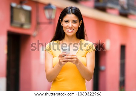 Happy young latin woman talking and texting on smart phone, wearing casual clothes sitting in with a take away coffee in a urban bench  Royalty-Free Stock Photo #1060322966