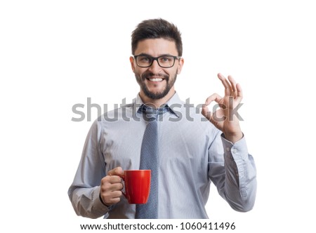 Young cheerful businessman in blue classic shirt smiling with wide opened mouth and holding red mug with drink. Portrait of successful entrepreneur showing ok sign against white background