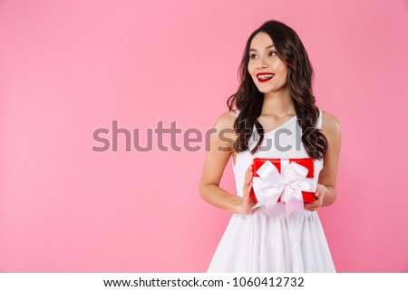 Image of cute pretty young asian woman standing isolated over pink background looking aside holding gift box.
