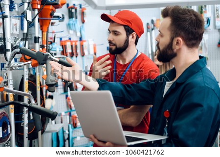 Two salesmen in red shirt and baseball cap and blue robe are checking tooks inventory with laptop in power tools store. Royalty-Free Stock Photo #1060421762