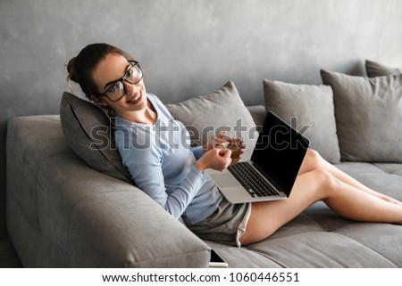 Portrait of a happy young woman drinking tea while sitting on a couch with blank screen laptop computer at home