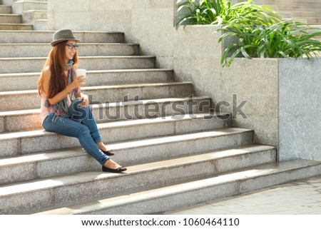 Taking short break from work: stylish Asian designer enjoying fragrant coffee while sitting on stairs illuminated with daylight, full length portrait