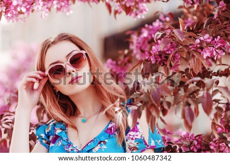 Outdoor close up portrait of young beautiful girl wearing stylish pink sunglasses, necklace, printed dress, posing in street, near spring blooming tree. Female fashion concept. Copy, empty space