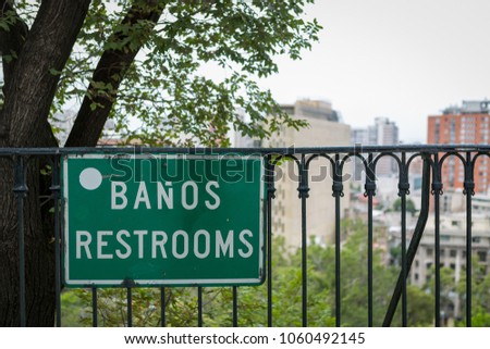 Bathroom sign in Spanish and English, white letter on green plate on a fence under a tree with a city view in a background.