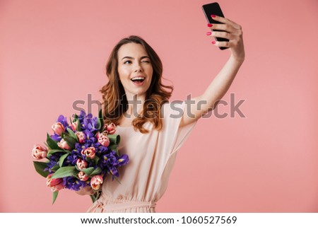 Portrait of a happy young girl in dress taking selfie with mobile phone while holding big bouquet of irises and tulips isolated over pink background