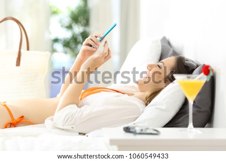 Side view portrait of a happy woman using a smart phone lying on a bed of an hotel room on summer vacations