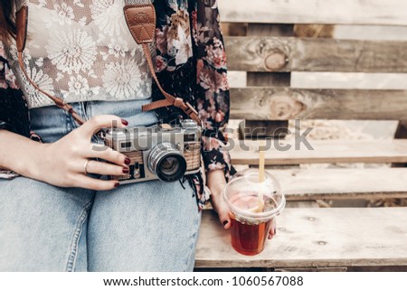 stylish hipster woman holding old photo camera and lemonade. boho girl in denim and bohemian clothes, sitting on wooden bench at street food festival. summer travel. space for text