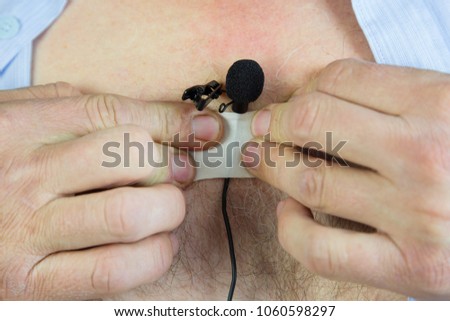 A man attaches a hidden microphone to his chest with an adhesive tape. Lavalier microphoneA private detective puts on a small microphone a buttonhole under his shirt for secret recording of the sound.