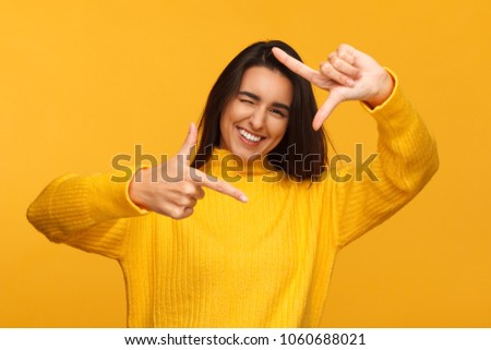 Cheerful young woman in yellow sweater winking and composing frame with hands. 