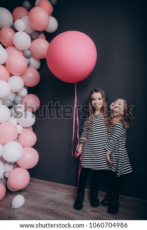 Two 4 years old girls in striped black and white dresses are having fun on a dark background with white and pink air balloons.
