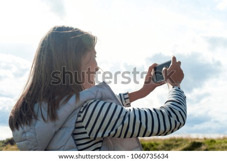 Cheerful young beautiful girl taking picture by smartphone in Scottish Highlands. Multicolored summertime horizontal outdoors image with cloudy sky background.