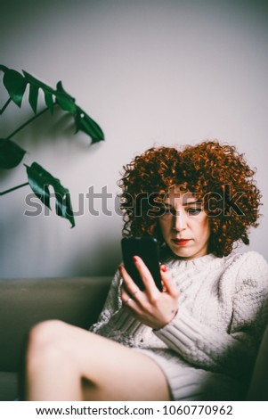 Redhead young woman with curly hair sitting and looking at smartphone screen on sofa (toned image, selective focus)