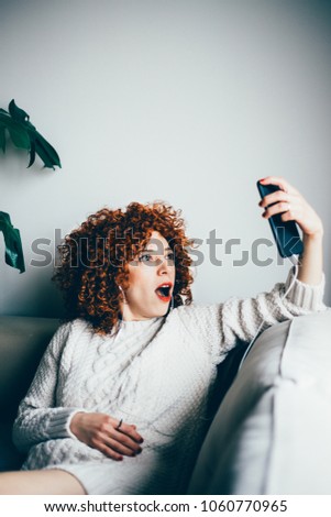 Surprised young woman with curly red hair, in earphones, sitting on sofa and communicating online by video chat (toned image, selective focus)