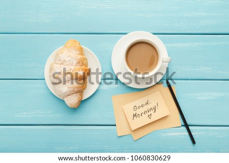 Coffee cup with croissant and note Good morning on blue rustic table from above. Top view on cozy and tasty breakfast, copy space