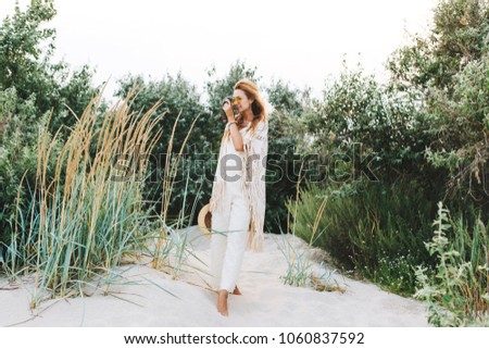 Full-length portrait of charming blonde photographer making photo of nature during walk in forest. Elegant woman with camera enjoying weekend and hobby spending time outdoor.