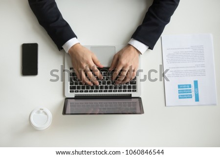 Top view of company CEO typing at laptop, handouts papers, coffee and smartphone around. Businessman working at office desk. Concept of hard work, flat lay, close up view Royalty-Free Stock Photo #1060846544