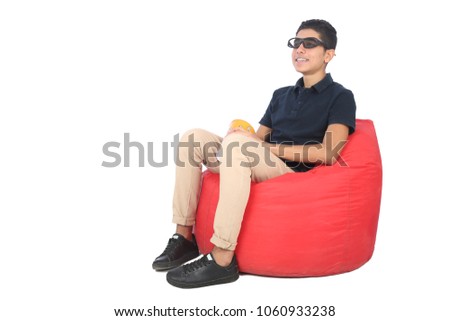Side shot  of a Teenager sitting on a bean bag wearing a 3D glasses and holding a popcorn cup in his hand eating it, isolated on white background