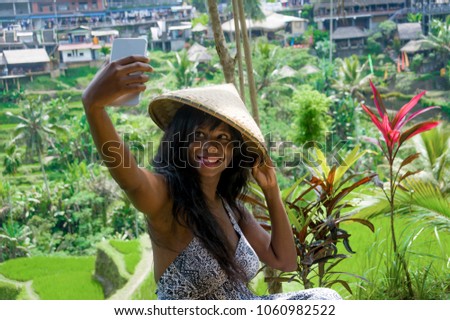 young attractive happy afro american black woman tourist taking selfie portrait photo with mobile phone camera while exploring rice fields forest and jungle in Bali wearing traditional Asian hat