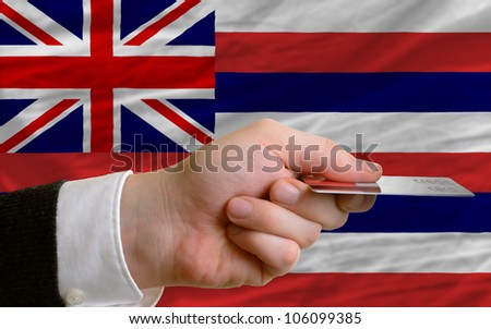 man stretching out credit card to buy goods in front of complete wavy national flag of american state of hawaii