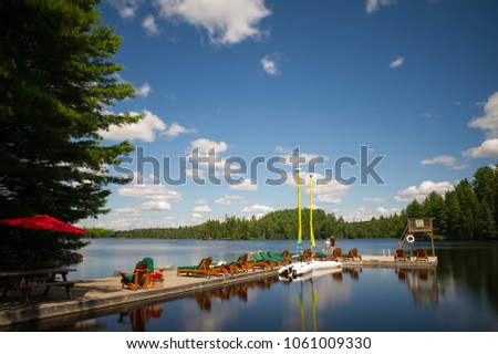 Adirondack and lounge chairs sitting on a wood dock facing a calm lake during a sunny day. Sailing boats are tied to the dock.