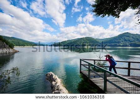 Woman photographer with a blue backpack on the lake Vidraru Carpathians Romania. Beautiful scenery of a sunny day: mountains, forests, lake, blue sky with clouds