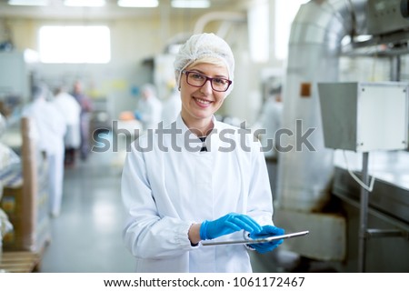 Young joyful beautiful female worker in sterile cloths holding a tablet and smiling for the camera near factory production line.