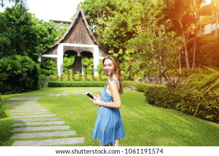 bright portrait of young woman holding phone, smiling and looking at camera on palms and house background with sunshine. Concept of travel, photo with copy space and new technologies