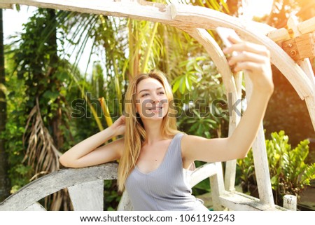 Young woman uses smart phone for selfie on sunny day, background of sunshine green palms in Thailand, Phuket. Concept of new technologies for better life, travel to tropical countries, remote work
