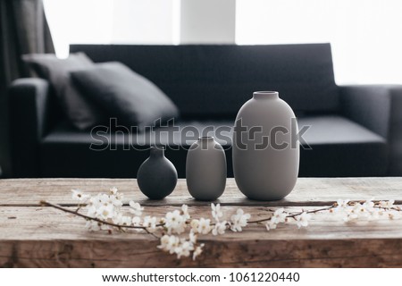 Minimalistic home decor on rustic coffee table over black sofa with cushions. Grey vases and spring flowers on wooden bench in small dark room interior. Scandinavian home style. Royalty-Free Stock Photo #1061220440