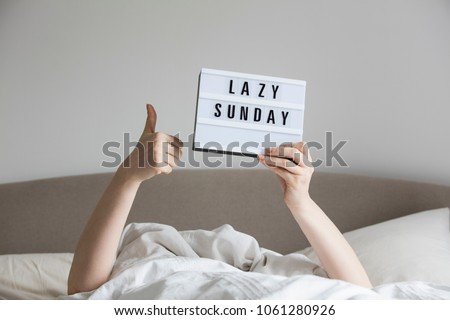 Female in bed under the sheets holding up a lazy sunday sign Royalty-Free Stock Photo #1061280926