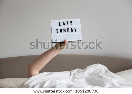 Female in bed under the sheets holding up a lazy sunday sign Royalty-Free Stock Photo #1061280938