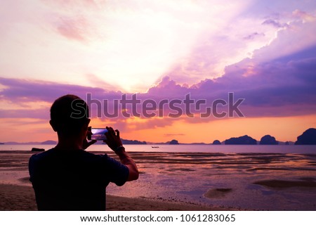 Silhouette of Asian middle aged man taking photos of sunset beach with smart phone. Summer, travel and technology concepts. Copy space. Back of young man taking a photo with phone in Krabi, Thailand.
