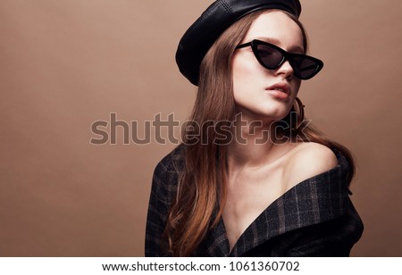 Fashion portrait of beautiful young woman in black leather beret cap, plaid jacket and cat eye retro sunglasses with massive golden earrings Royalty-Free Stock Photo #1061360702