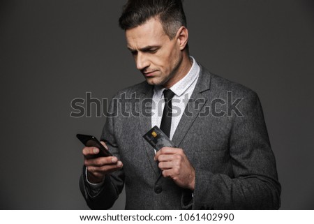 Photo of confident entrepreneur man in suit and tie holding smartphone and credit card for paying online isolated over gray background