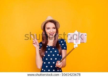 Front view portrait of pretty, cheerful, confident, successful girl  holding, using selfish stick with smart phone, making selfie, gesture v-sign to the front camera, standing over yellow background