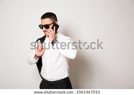 Young brunette guy with beard. Good smile with white teeth. Wonderful, emotional. In black pants and white shirt. The jacket on the shoulder. On a white background. With the phone in hand.