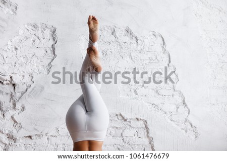 Concentration and preparation. Fit, unidentified young woman in sports suit makes stand on  head with her legs crossed - yoga exercise in an empty gym with white wall background. Copy space Royalty-Free Stock Photo #1061476679