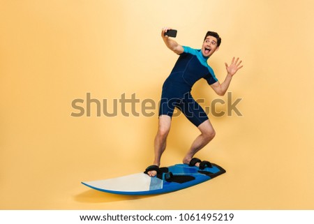 Full length portrait of an excited young man dressed in swimsuit taking a selfie while surfing on a board isolated over yellow background