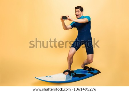 Full length portrait of a confident young man dressed in swimsuit talking a picture with mobile phone while surfing on a board isolated over yellow background