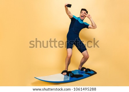 Full length portrait of a smiling young man dressed in swimsuit taking a selfie while surfing on a board isolated over yellow background