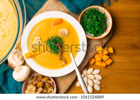 hot pumpkin soup in glass pan and in white plate, decorated with mini carrot, pumpkin seeds, mushroom, honey agaric and green kelp on wooden board. Vegan.