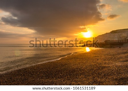 Glorious sunset on the beach, with vast storm cloud in the sky. The golden sun sinking behind the cliffs on the pebble beach. The smooth sea calmly lapping the shore. The sun reflecting in the water.