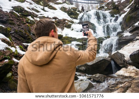 A young man takes a picture of a waterfall on a mobile phone in the spring in the mountains
